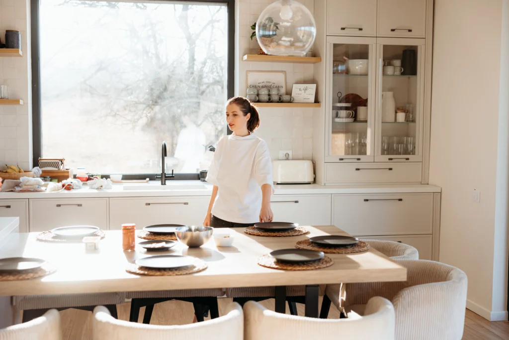 Woman setting a wooden table in a sunlit, modern kitchen with open shelves and neutral cabinetry, designed for functionality and aesthetics.
